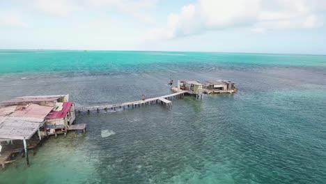 Drone-shot-man-jump-on-old-wooden-pier-from-Palafito-stilt-house,-behind-view,-Los-Roques