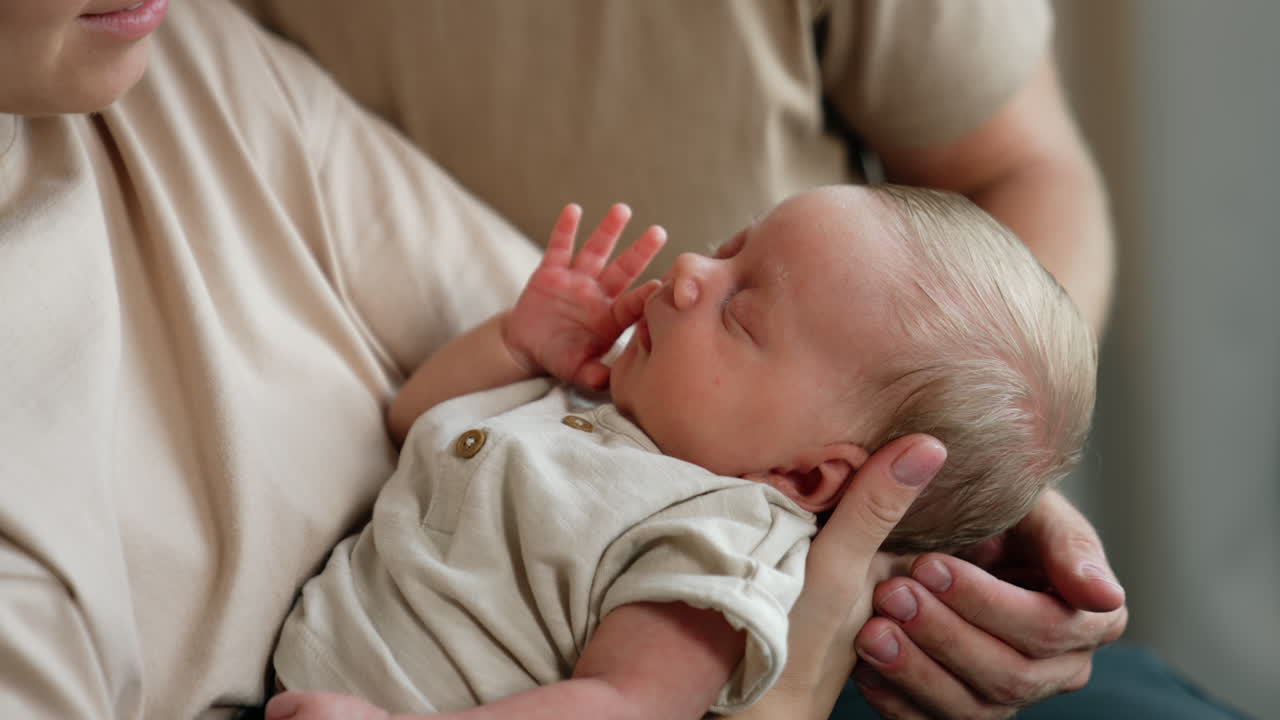 Adorable tiny newborn sleeping in mom's hands. Careful dad's hand strokes little baby's head. Close up.
