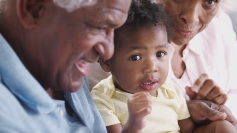 grandparents sitting on sofa at home playing game with baby granddaughters
