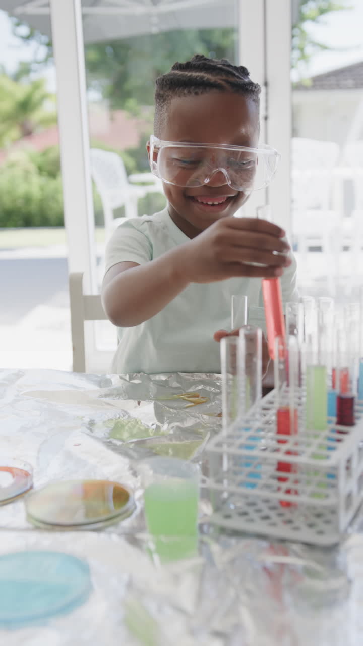 Vertical video of african american boy sitting at table holding test tubes with liquid, slow motion