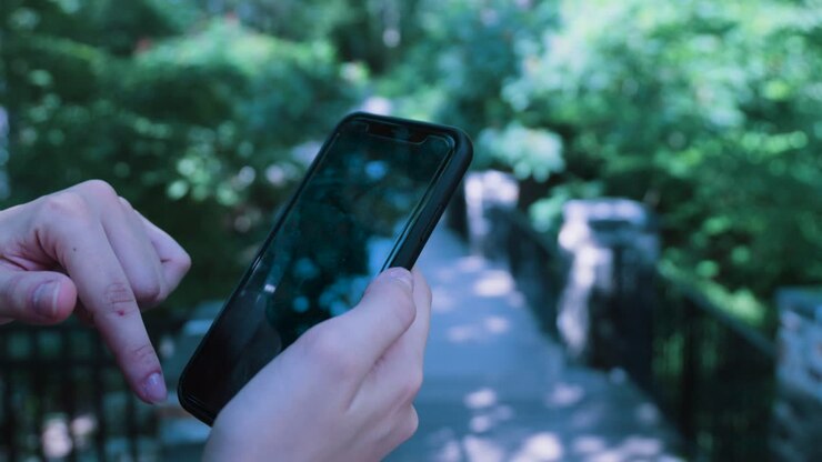Una mano scorre su un iPhone nero sullo sfondo di un sentiero di parco alberato e verdeggiante in una giornata di sole.