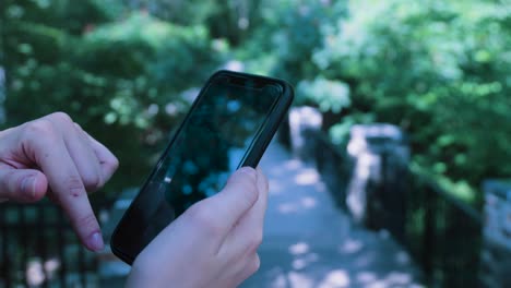 a hand scrolls through a black iphone with the backgrop of a forested green-lined park pathway on a sunny day