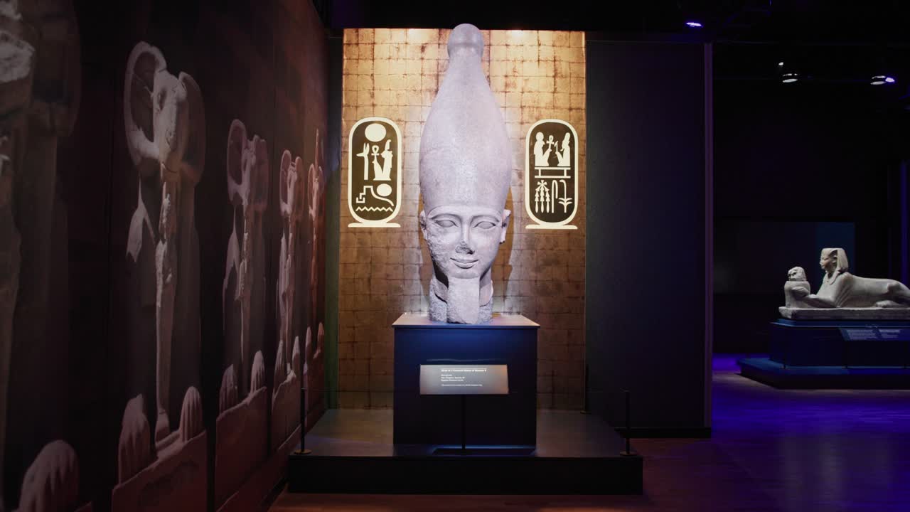 Veduta frontale di una statua del Faraone Ramses II esposta in una galleria museale scarsamente illuminata, illuminata contro uno sfondo testurizzato con simboli geroglifici.