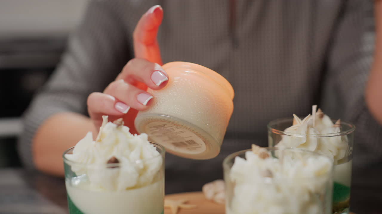 Creative lifts glowing candle vessel placed among beautifully decorated desserts topped with white floral designs, creating warm elegant scene blending sweetness, creativity, and cozy atmosphere