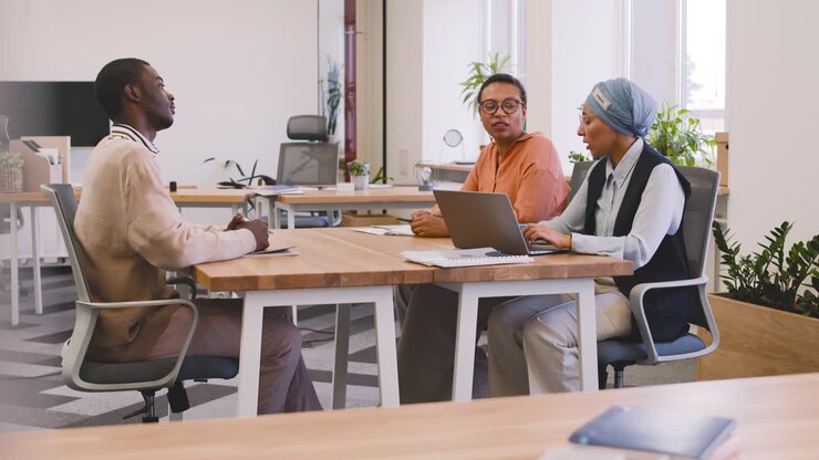 An Woman And A Muslim Woman Co Workers Interview A Young Man Sitting At A Table In The Office 16