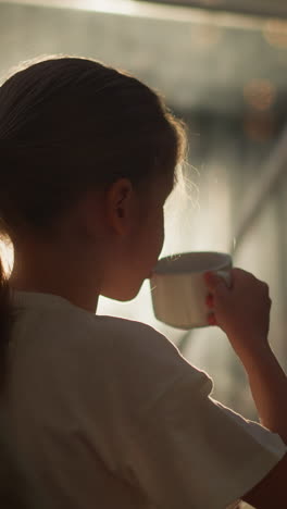 child drinks from cup in morning. girl enjoys warmth of favorite beverage sitting in room against early rays of sunlight. cozy morning at home