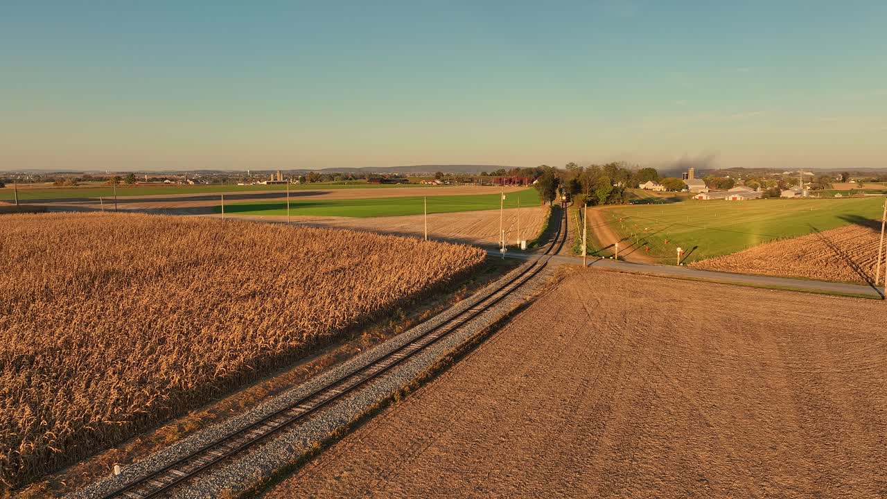 In the distance a steam train moves along the tracks surrounded by golden cornfields and lush green pastures under a clear blue sky. Farms and rural life are visible in the distance.