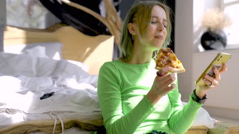 young blonde woman watching something funny on mobile phone and eating pizza while sitting on the floor at home