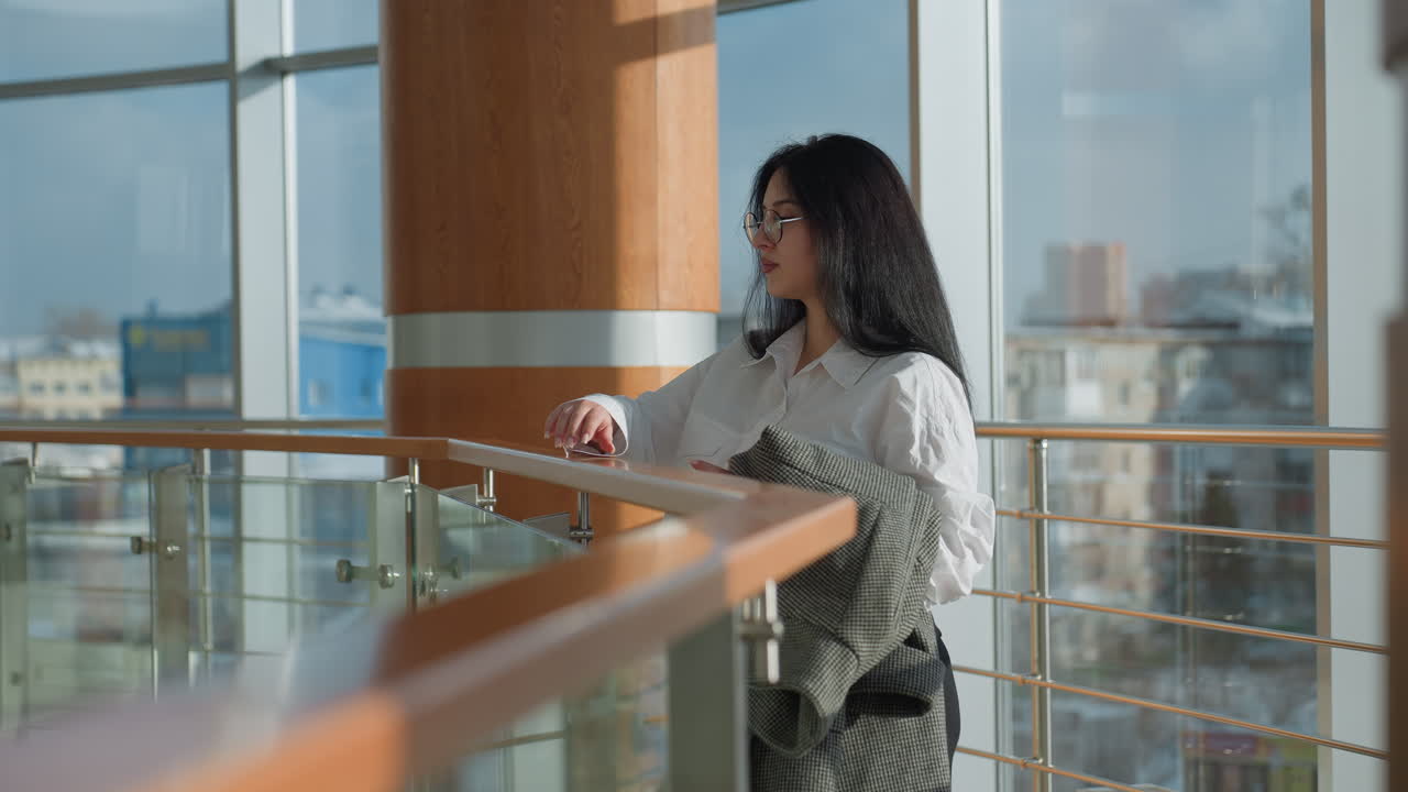Elegant young student in white shirt holding checkered coat walks to wooden frame railing inside sunlit mall, gently rests on it while looking down through large glass window with thoughtful gaze