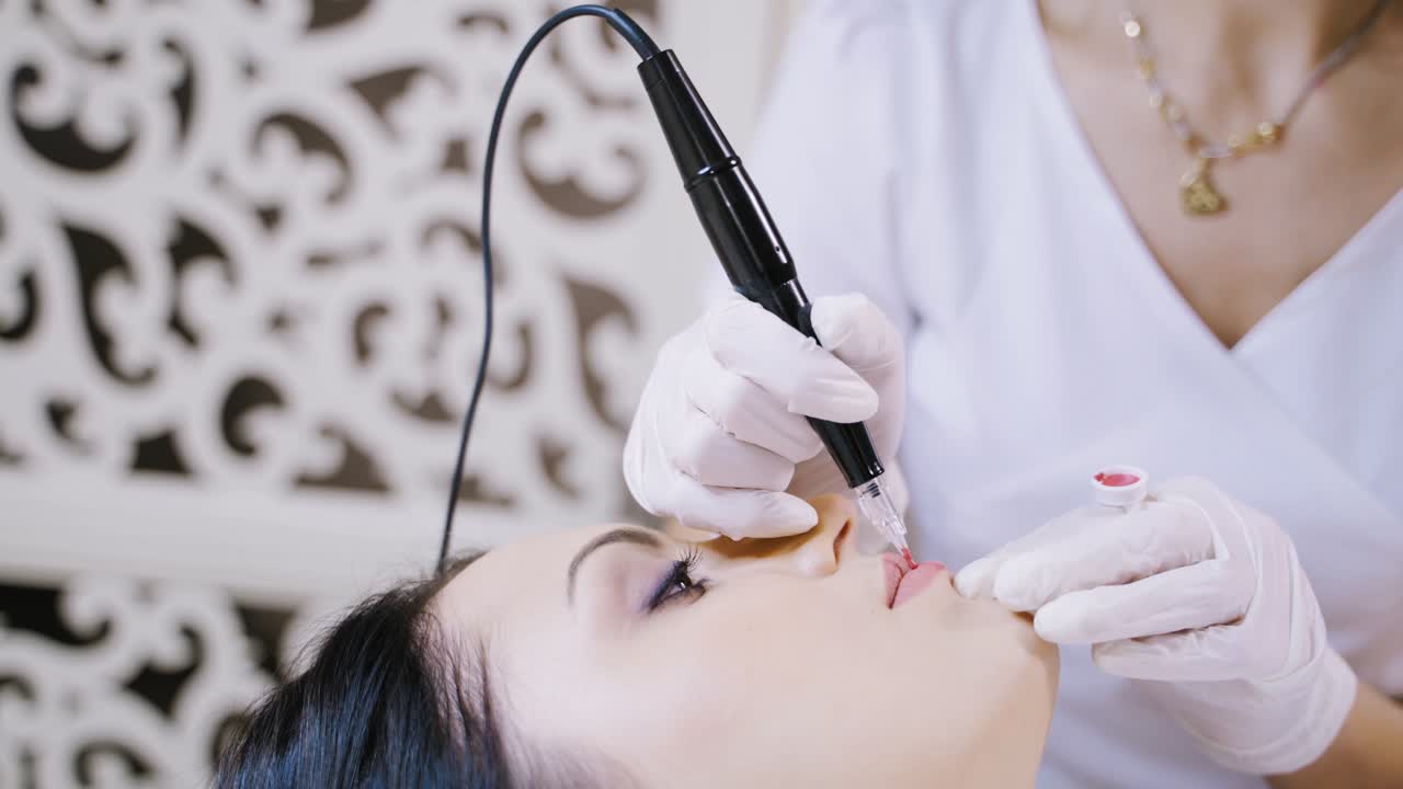 Skilled cosmetician applying permanent make-up on lips in a beauty salon. Young woman undergoing procedure of permanent lip makeup with tattooer machine. Close-up