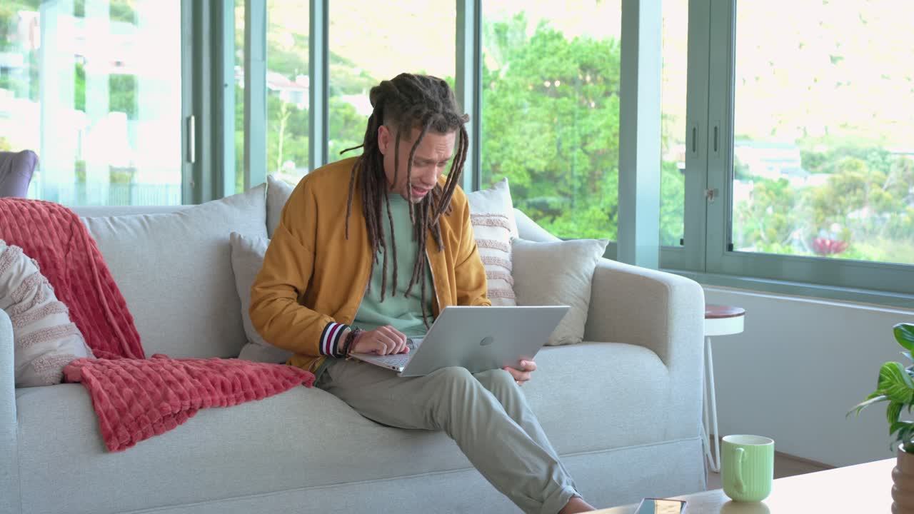 Entering living room, man in coat sitting on sofa grabbing phone, opening laptop for remote work