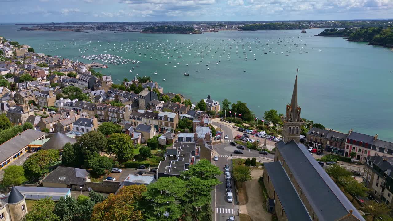 Dinard coastal town panorama with historic buildings and Church of Our Lady of the Emerald, boats on the Rance estuary and distant Saint Malo under a bright summer sky, drone pulling away