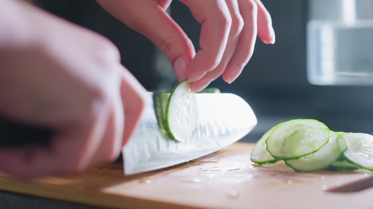 Close-up of hand slicing cucumber with knife on wooden cutting board in bright kitchen setting, focus on fresh, healthy ingredient preparation in a well-lit culinary environment