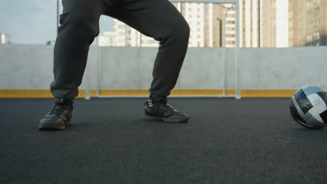 lower angle view of athlete in squatting position with hands clasped, performing intense workout on outdoor sports ground, as soccer ball rolls nearby