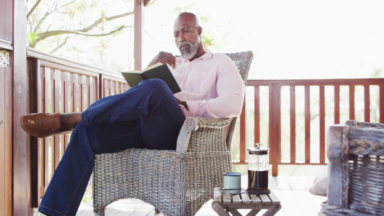 Happy senior african american man spending time in log cabin, reading book on balcony, slow motion