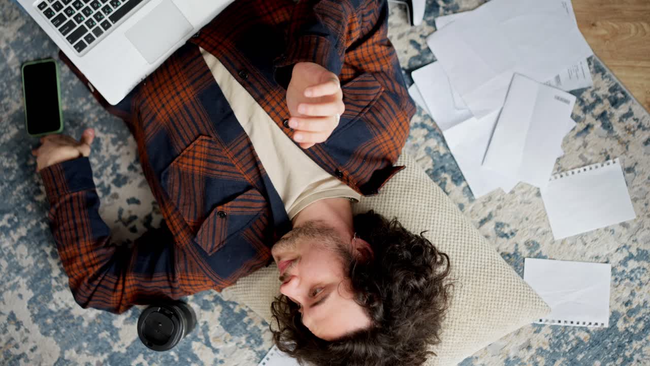Camera rotation Tired brunette guy with curly hair lies on the floor on a pillow around him sheets of paper coffee laptop and smartphone at home in a modern apartment