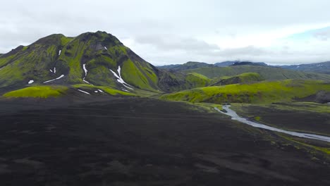 aerial drone footage flying and gliding over dark colored rocky and green moss covered iceland landscapes with large mountains in the background and a small narrow river flowing on the right hand side