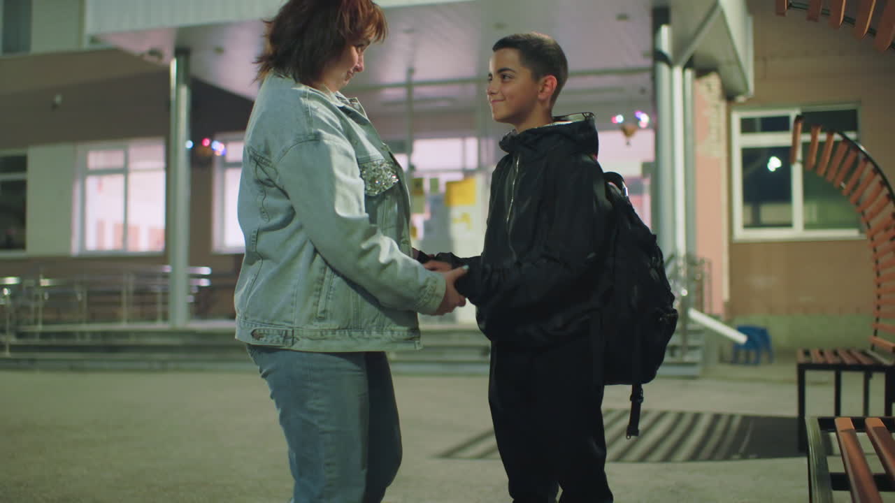Woman adjusts jacket of boy standing near school entrance during early morning, showing care and attention before start of day, with soft lighting and empty surroundings conveying peaceful moment