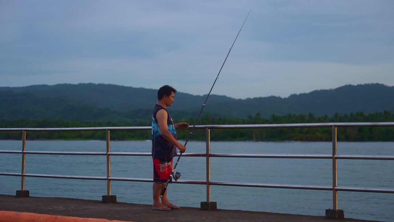 Sebuah bidikan tetap seorang pria dengan kaus tanpa lengan dan celana pendek menyandarkan tongkat pancing di pagar logam di sepanjang jalan tepi laut dengan latar belakang alam di Pelabuhan Mauban, Provinsi Quezon, Filipina