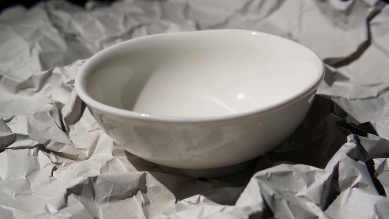 Shifting camera revealing white ceramic bowl on crumpled paper, zooming in to show glossy rim