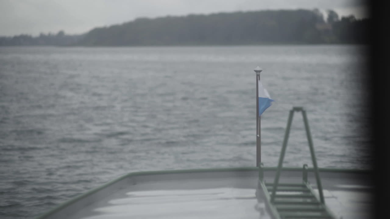 A white and blue flag waving in the wind on a cruising boattransporting people at the beautiful lake chiemsee during acloudy summer morning