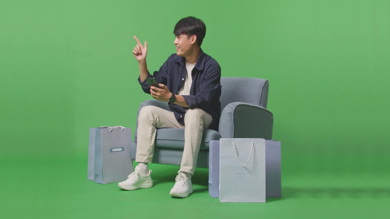 휴대 전화 를 사용 하고 손가락 을  으로 가리키며 녹색 스크린 앞 에 소파 에 앉아 있는 동안 카메라 를 향해 미소 짓는 아시아인 쇼핑 남자