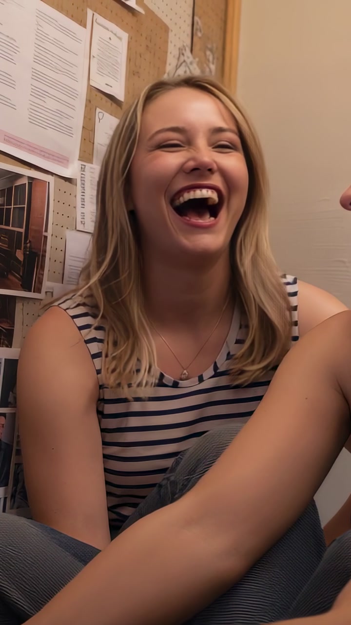 Vertical video: Laughing woman in striped top reacting to friend chat in dorm room, with corkboard