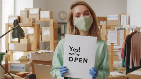 woman in mask and gloves holding sign saying "yes! we're open" in a retail store