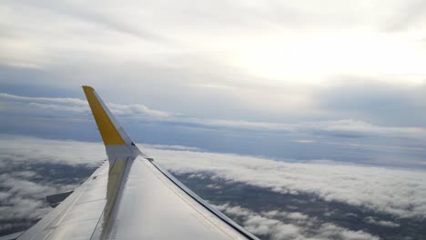 pov airplane passenger window: white clouds below with view of aircraft wing in flight with bright white sun in sky, handheld slow motion