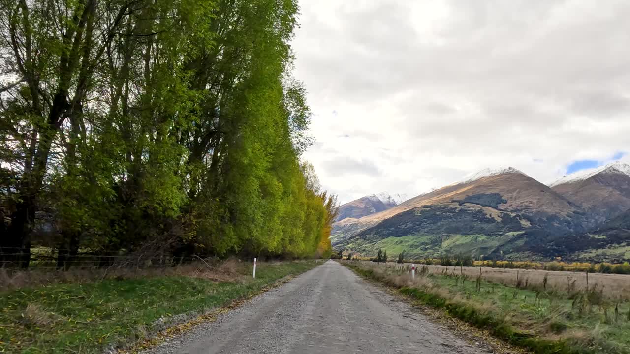 Forward-moving vehicle view along rural gravel road, autumn trees, overcast sky, distant mountains visible