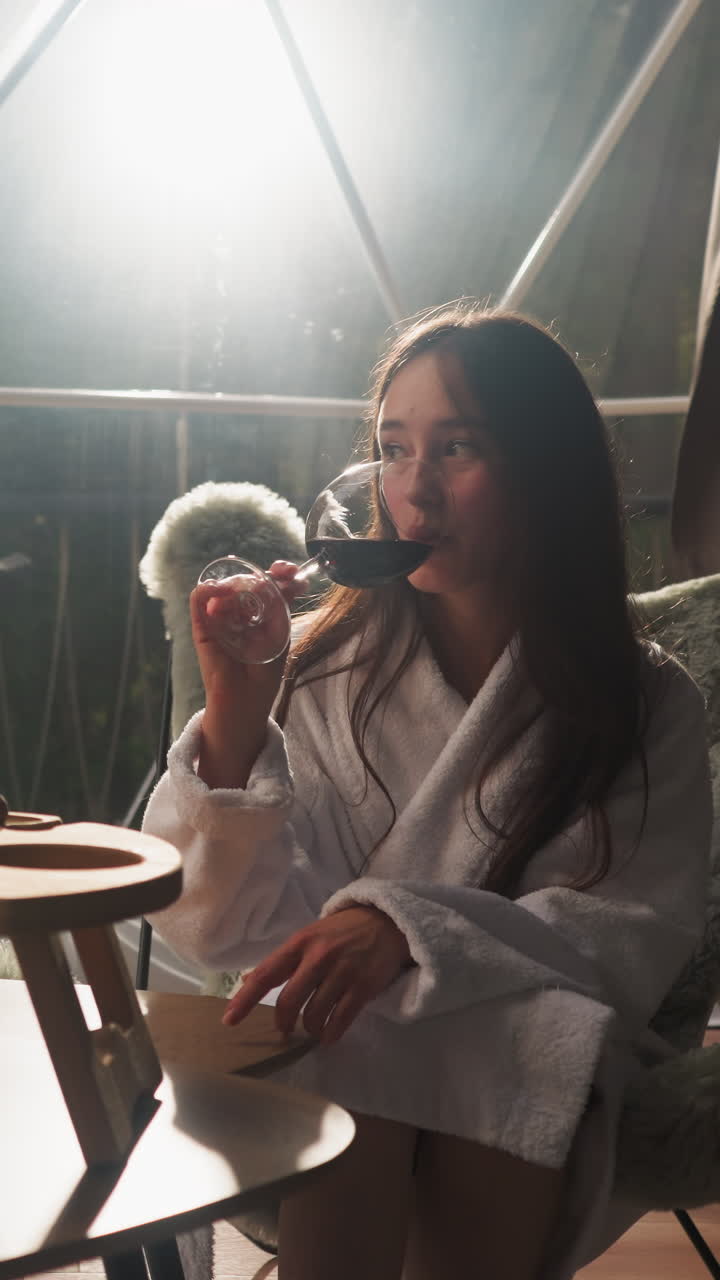 Woman tastes glass of wine at glamping house. Newlyweds in soft bathrobes enjoy romantic evening with delicacy snacks and beverage on glass terrace