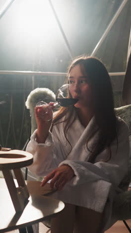 woman tastes glass of wine at glamping house. newlyweds in soft bathrobes enjoy romantic evening with delicacy snacks and beverage on glass terrace