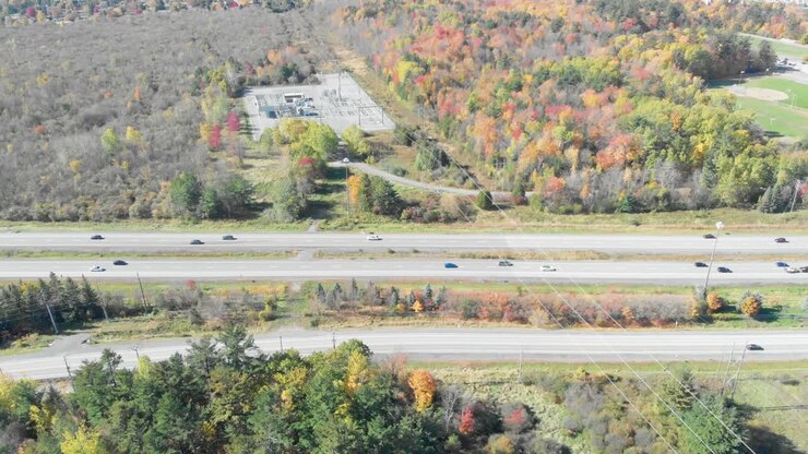Aerial shot to the left of a large 4 lane highway with lots of cars going each way in between two autumn coloured forests with hydro lines going over the lanes