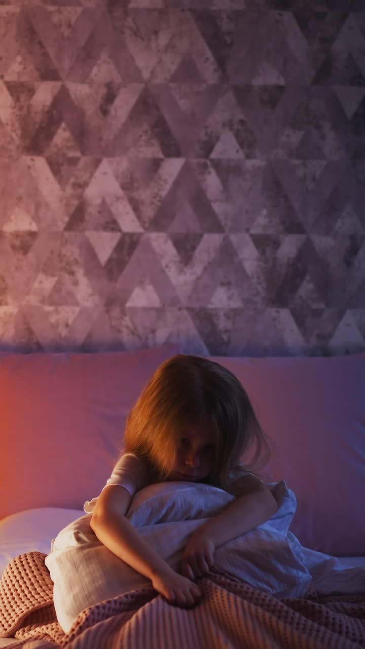 Nervous little girl scratches pink plaid holding soft pillow on bed near illuminated by night lamp play wigwam in semidark bedroom at home slow motion