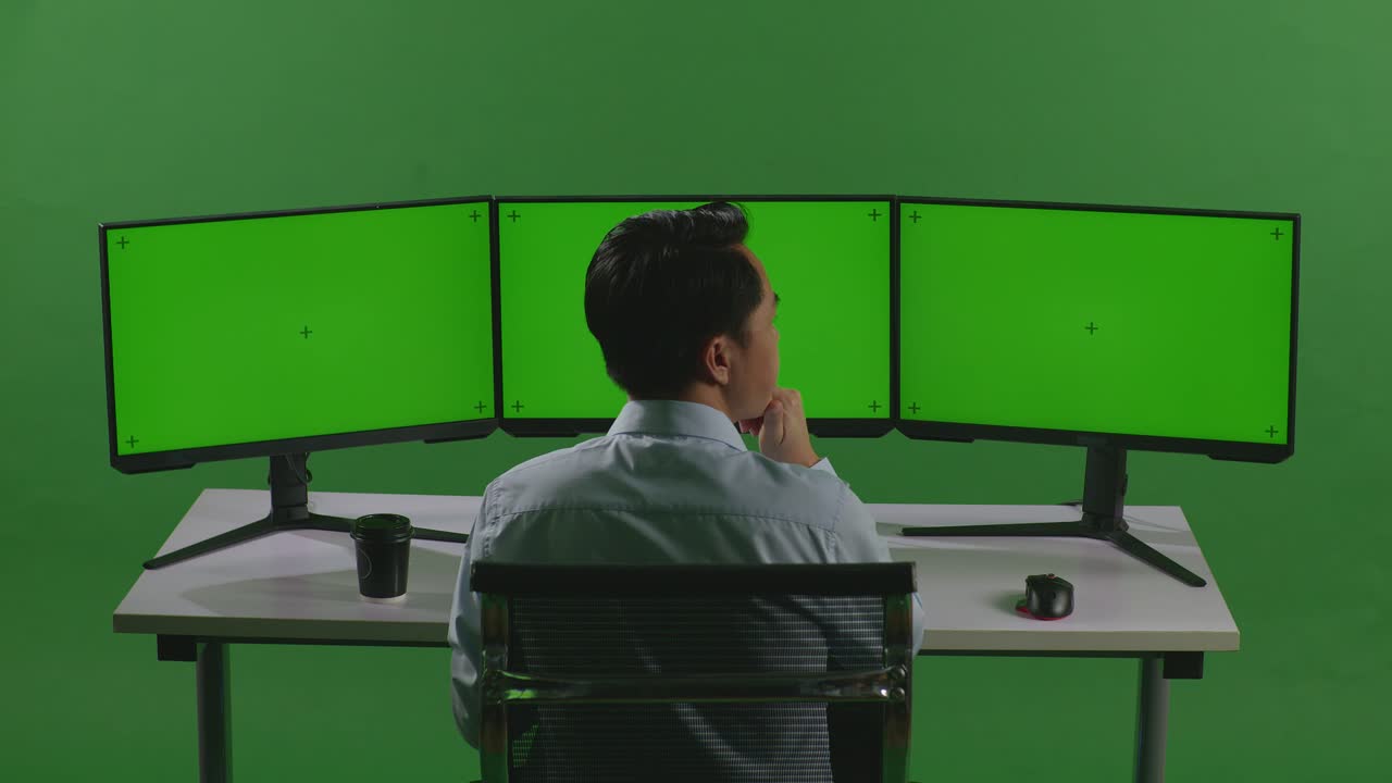 녹색 스크린 스튜디오에서 모의 여러 컴퓨터 모니터와 함께 작업하는 동안 무언가에 대해 생각하는 아시아 남자의 뒷면