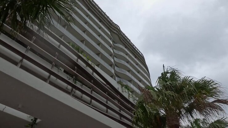Vista en ángulo contrapicado con paneo pasando por palmeras tropicales y arquitectura moderna curva bajo cielos nublados