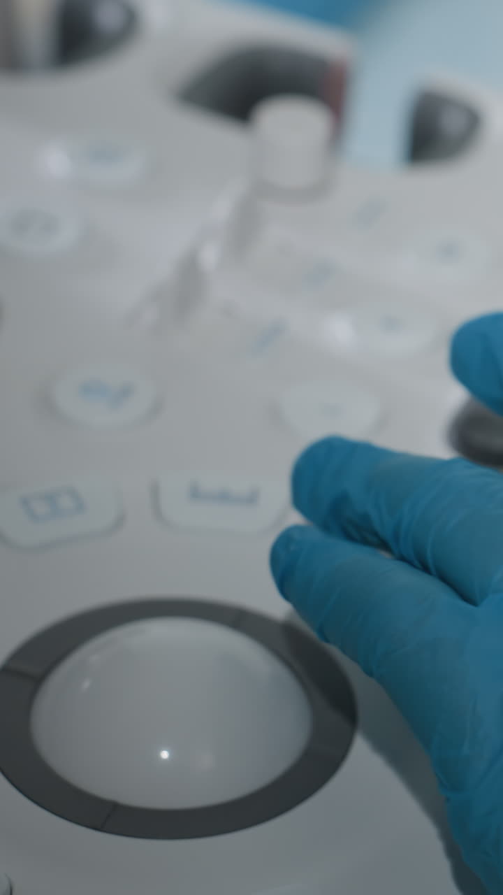 Mano enguantada sobre la consola de ultrasonido, ecografista ajusta los controles con un guante de nitrilo azul, primer plano de la trackball y los botones, entorno hospitalario clínico, técnica estéril e higiene, control de precisión