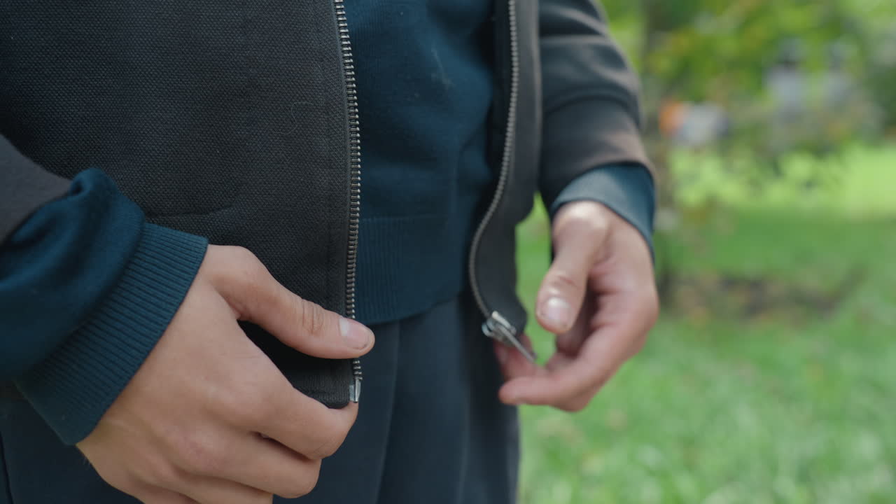 Seseorang mengancingkan mantel di luar ruangan, tampilan dekat tangan yang mengamankan mantel di lingkungan taman yang indah, bidikan detail seseorang yang menutup resleting jaket dengan kain bertekstur di bawah sinar matahari lembut di lingkungan taman