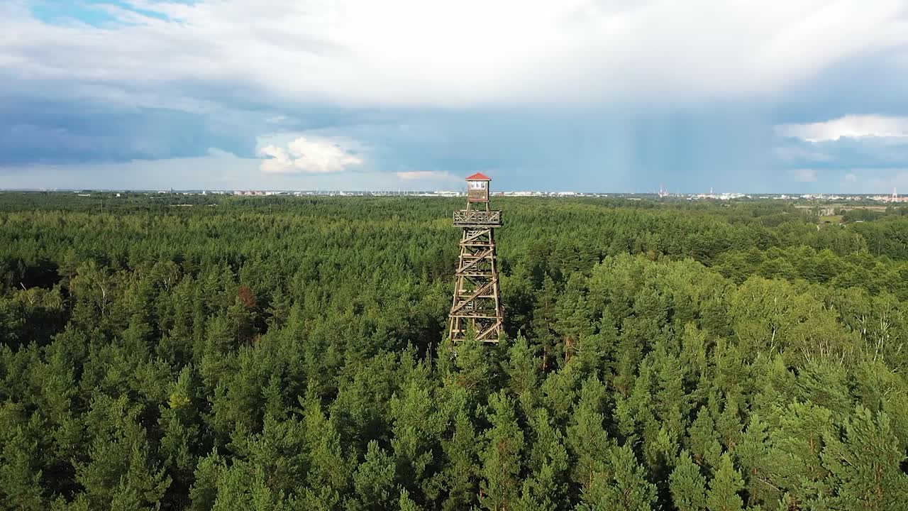 A drone orbits a watchtower enveloped by lush forest, while dramatic dark blue skies signal an oncoming storm and sunlight streams create dazzling color plays through the clouds