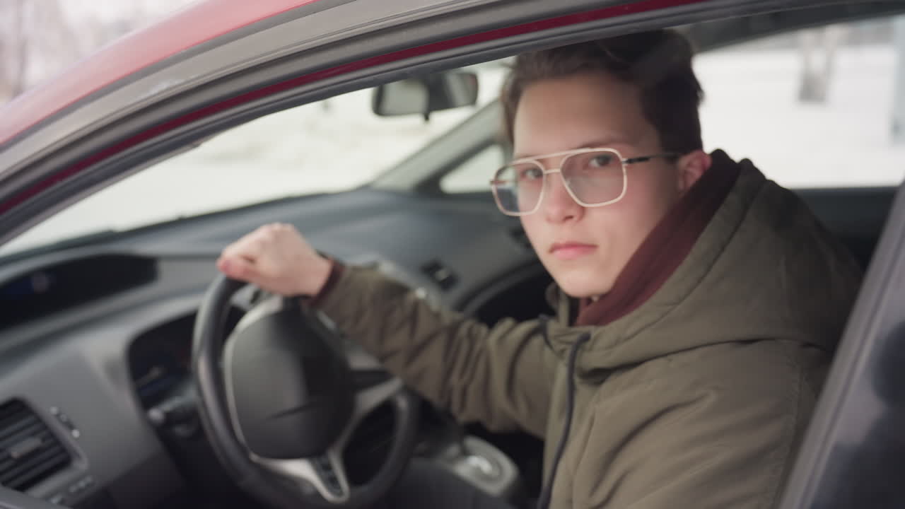 boy in sunglasses inside car lowers driver window and gazes through raindrop-covered glass toward camera with cold snowy winter street visible in background during quiet moment