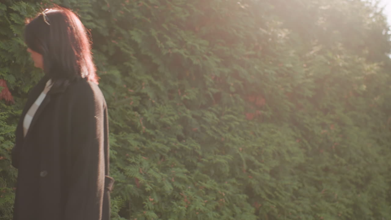 겨울 코트를 입은 패셔너블한 소녀가 공원 산책로를 따라 걷고, 손은 초록색 울타리 옆 가을 단풍 위를 스치듯 지나가고, 햇살이 나무 사이로 부드럽게 비치는 따뜻한 햇살 아래 야외 산책을 즐기는 모습이에요.