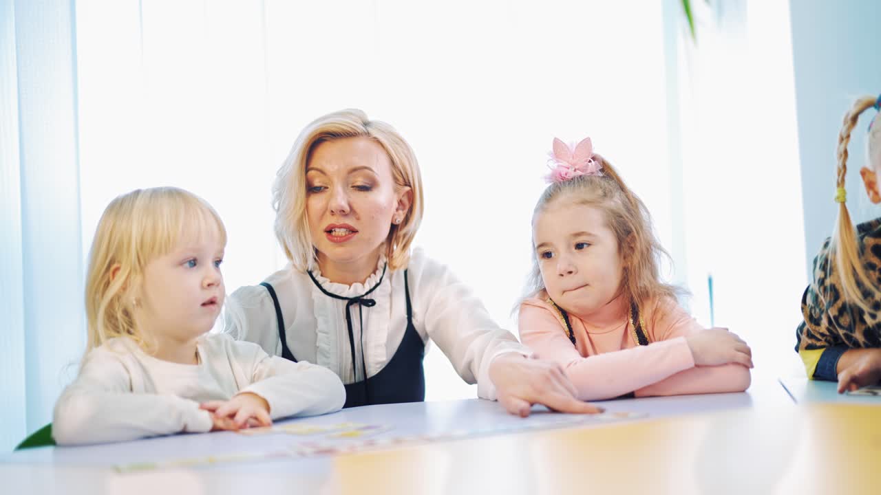 Teacher teaching sitting close to little kids. Small girls listen to their teacher in the classroom. Woman with children at the table.