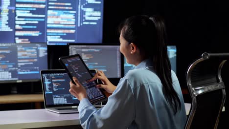 back view of asian female programmer looking at database on tablet while writing code by a laptop using multiple monitors showing database on desktops in the office