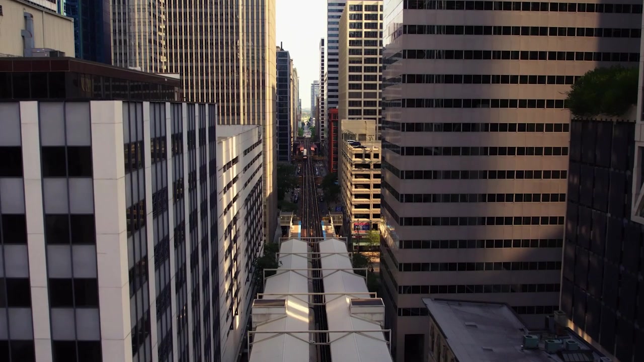 Vista drone che inquadra dall'alto un corridoio ferroviario nel centro di Chicago con una stazione CTA e binari sopraelevati, incorniciato da densi grattacieli