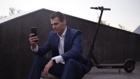 Attractive-Businessman-Sitting-On-The-Stairs-And-Does-Video-Cahtting-On-Smartphone-Outside