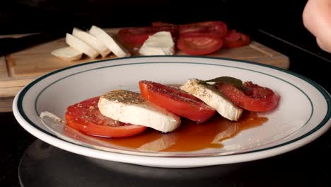 Eine-Weiße-Männliche-Hand-Legt-Zwei-Pfefferminzblätter-Auf-Einen-Tomaten-Mozzarella-Salat.-Im-Hintergrund-Sind-Ein-Messer-Und-Ein-Holzschneidebrett-Zu-Sehen
