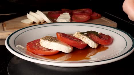 a white male hand puts two peppermint leaves on a tomato and mozzarella salad in the background is a knife and a wooden cutting board