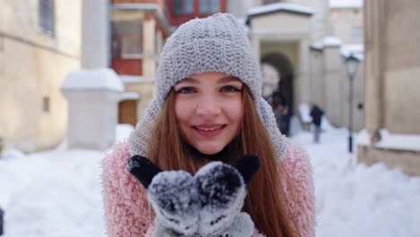 a young woman smiles and holds snow in her gloved hands in a snowy european city.