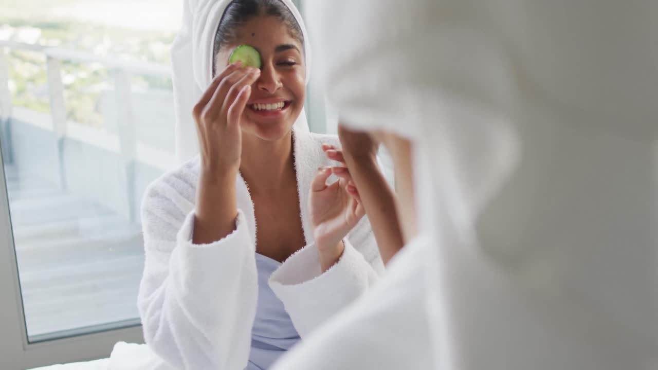 Video of happy diverse female friends in robes having fun with cucumber slices