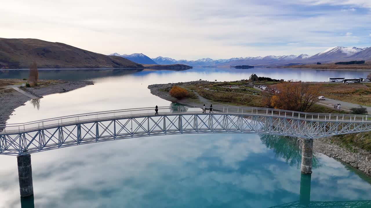 Drone footage captures tourists walking across a bridge over Lake Tekapo, surrounded by clear blue water and mountainous landscape
